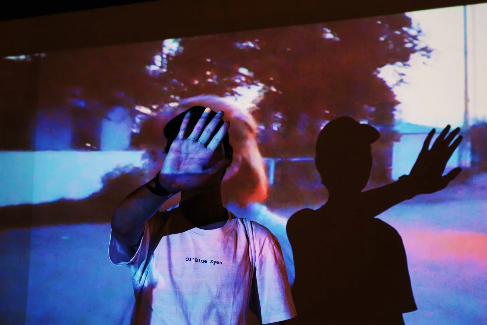 A man holds a hand up to his face while his shadow reaches out beside him on a projected wall